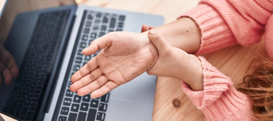 mulher com os braços sobre uma mesa de trabalho com notebook sentindo dor nos punhos